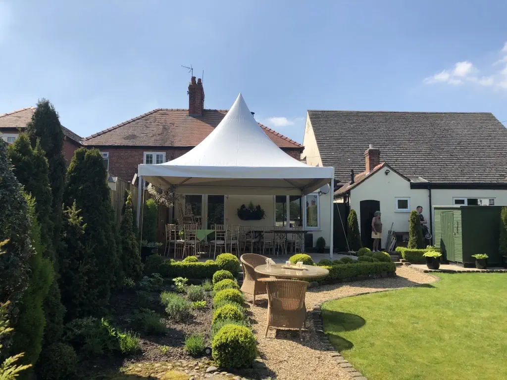 A landscaped backyard with a patio area covered by a white canopy, resembling elegant marquees. There are wicker furniture and a small path leading to a lawn. Two people stand near a shed and a house in the background, creating an inviting outdoor retreat.